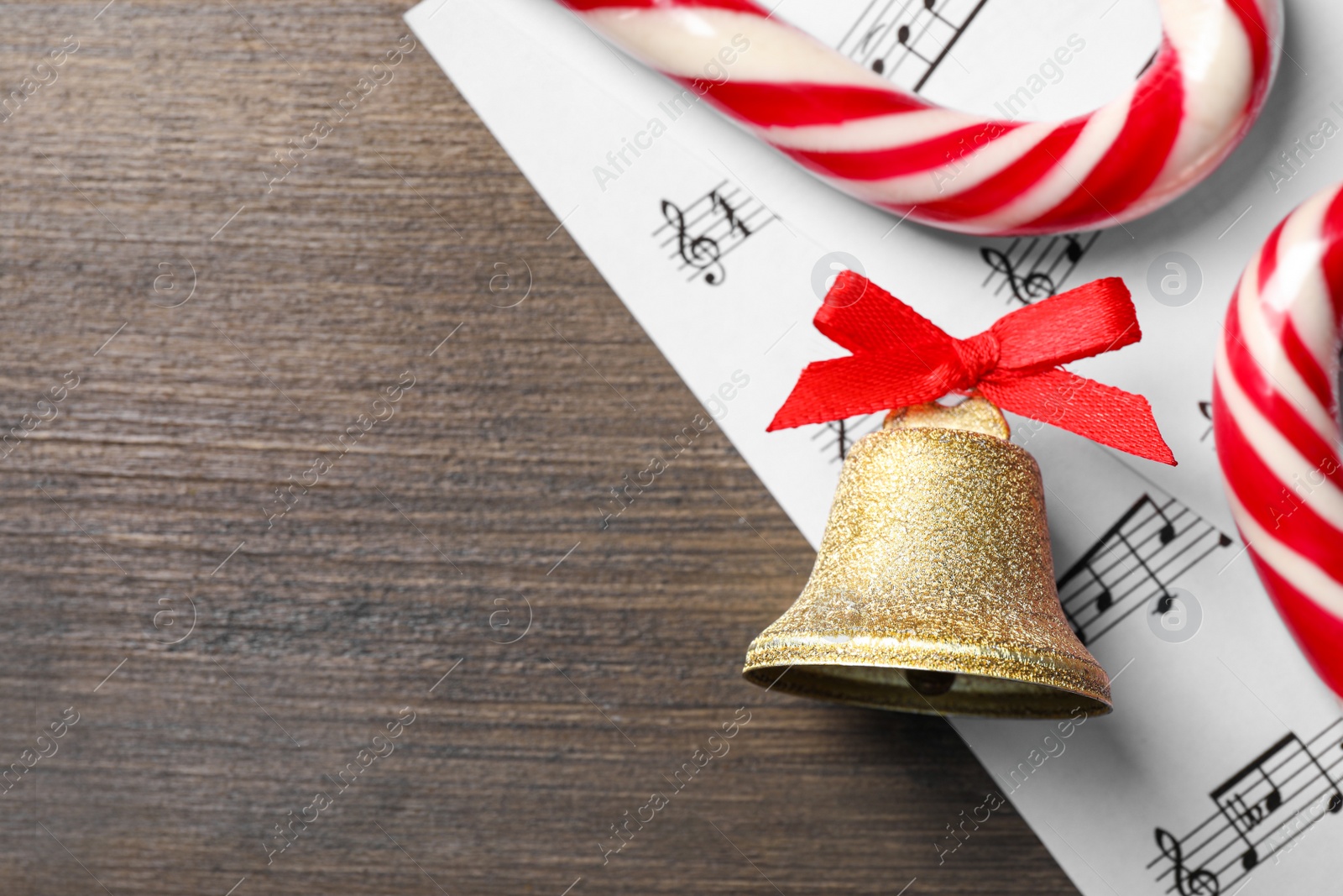 Photo of Golden shiny bell with red bow, music sheets and candy canes on wooden table, flat lay. Space for text. Christmas decoration