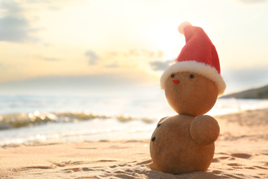Photo of Snowman made of sand with Santa hat on beach near sea, space for text. Christmas vacation