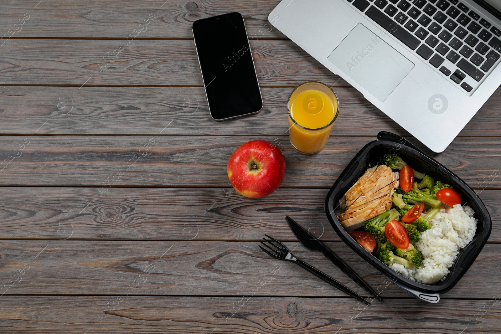 Photo of Container of tasty food, glass of juice, laptop, apple and smartphone on wooden table, flat lay with space for text. Business lunch