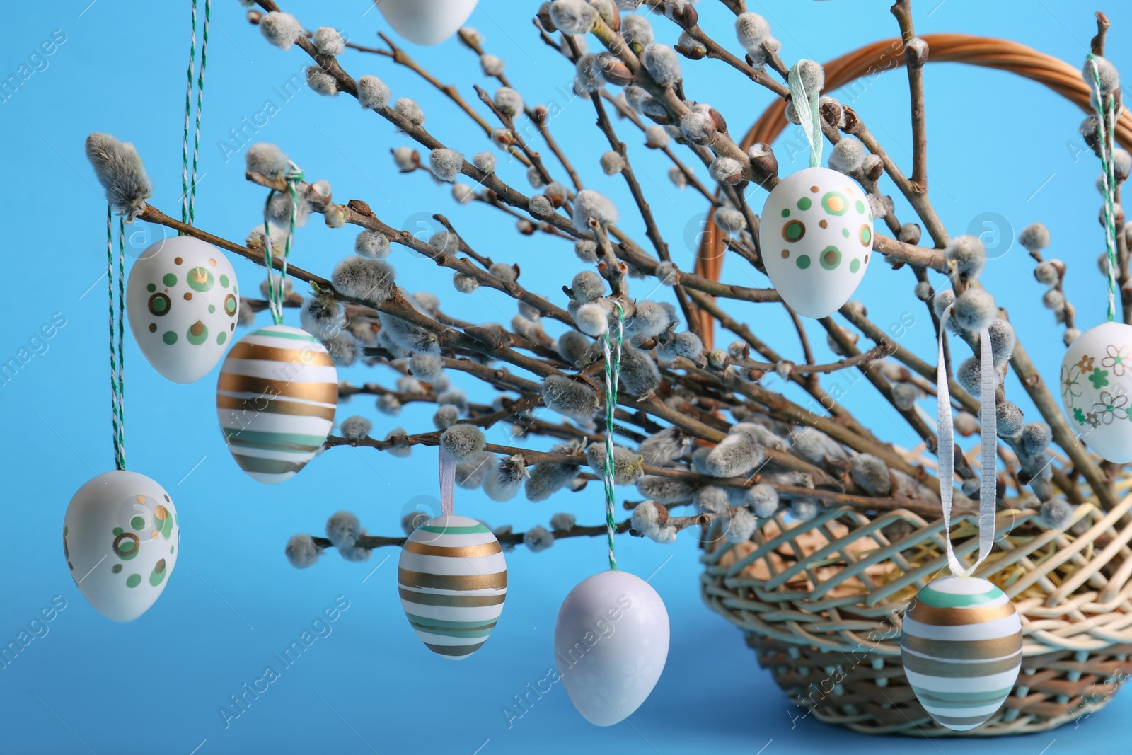 Photo of Wicker basket with beautiful willow branches and painted eggs on light blue background, closeup. Easter decor