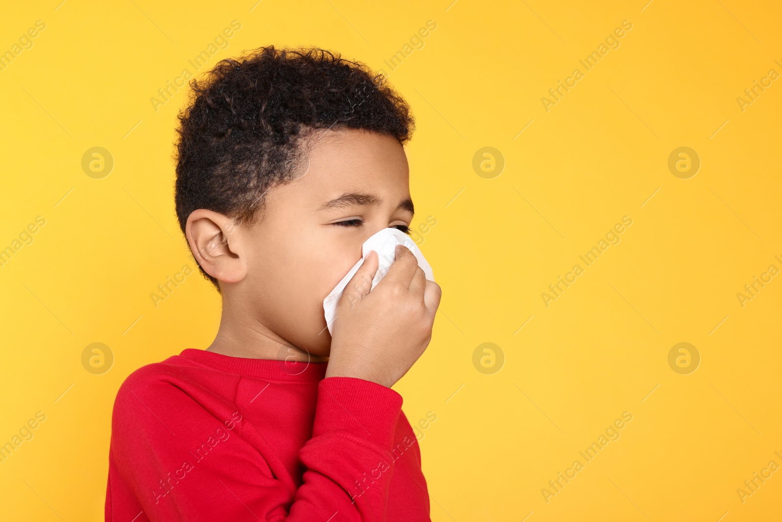 Photo of African-American boy blowing nose in tissue on yellow background, space for text. Cold symptoms