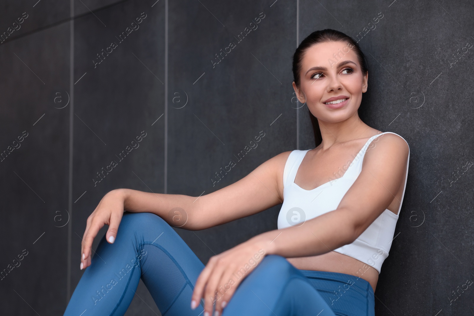 Photo of Smiling woman in sportswear sitting near dark grey wall