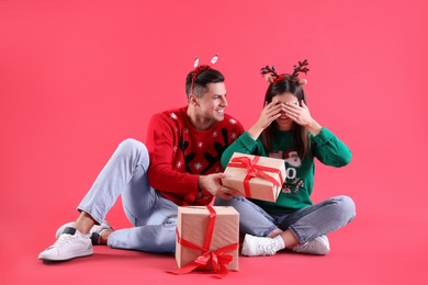 Photo of Beautiful happy couple in Christmas headbands and sweaters sitting with gifts on red background