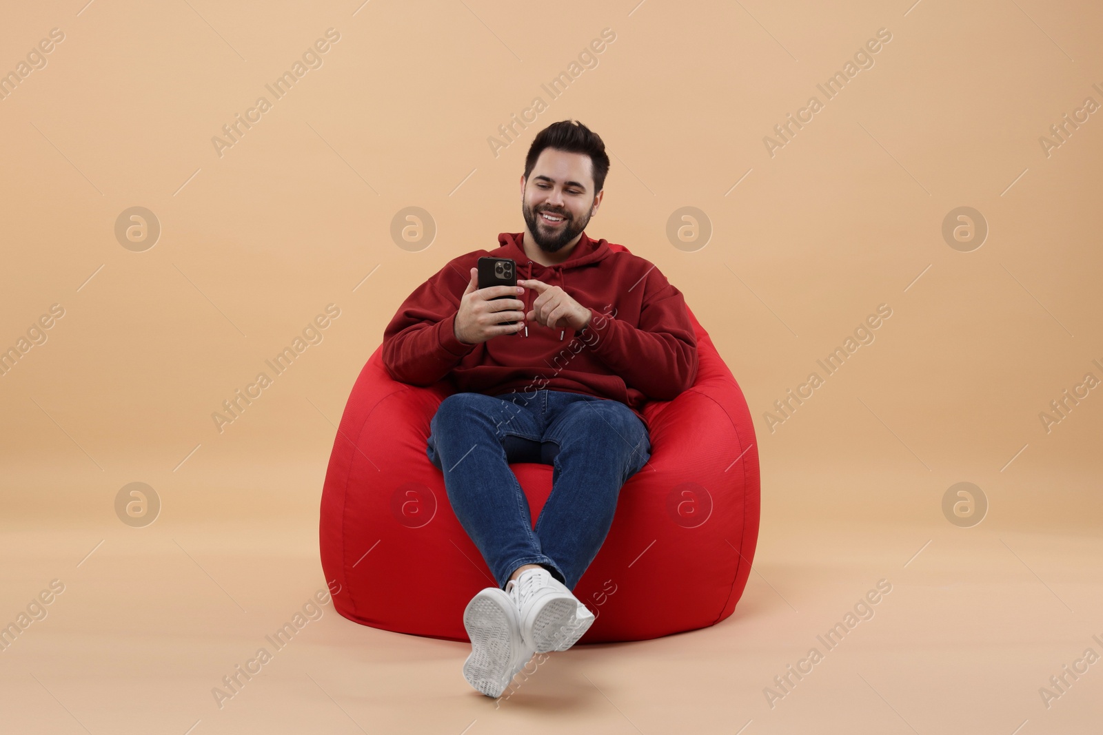 Photo of Happy young man using smartphone on bean bag chair against beige background