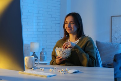 Photo of Beautiful young woman eating popcorn while watching film on computer at table indoors at night