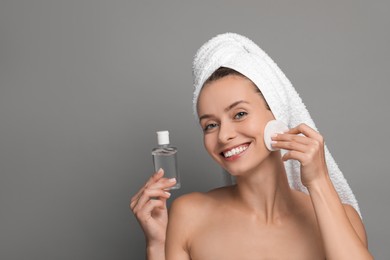 Smiling woman removing makeup with cotton pad and holding bottle on grey background. Space for text