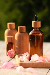 Bottles of rose essential oil and flowers on white wooden table outdoors, closeup