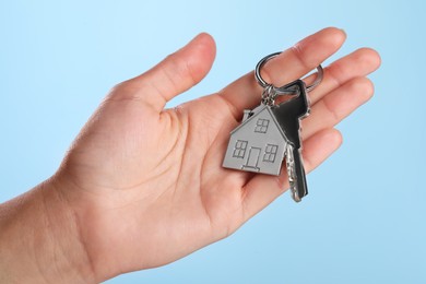 Woman holding key with metallic keychain in shape of house on light blue background, closeup