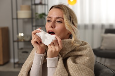 Photo of Sick woman wrapped in blanket with tissue sneezing at home. Cold symptoms