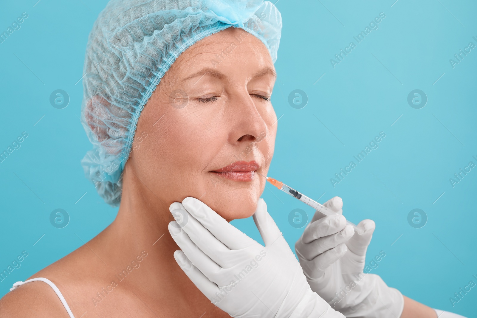 Photo of Doctor giving lips injection to senior woman on light blue background. Cosmetic surgery