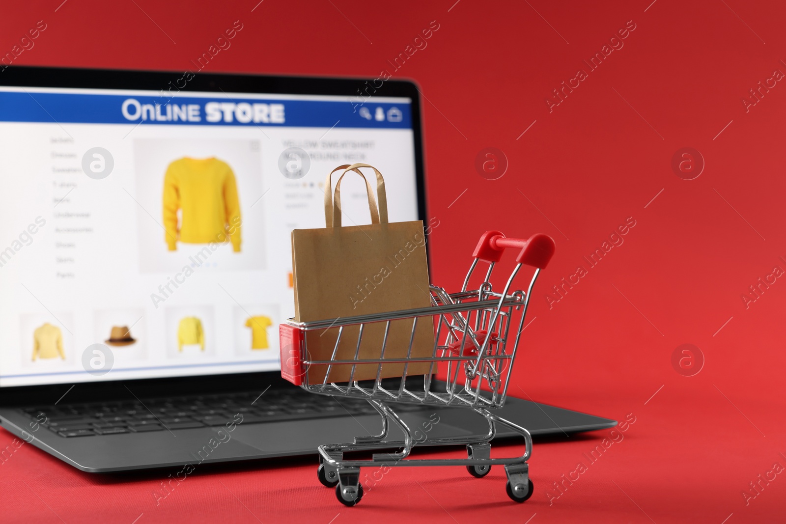 Photo of Shopping bag in mini cart and laptop on red background, selective focus