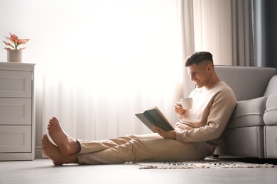 Photo of Man with cup of hot drink reading book at home. Floor heating concept