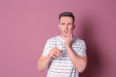 Young man with sensitive teeth and glass of cold water on color background