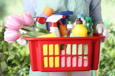 Photo of Woman holding basket with spring flowers and cleaning supplies on green blurred background, closeup