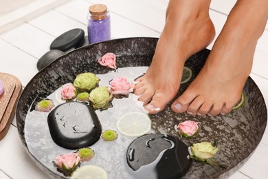 Photo of Woman soaking her feet in plate with water, stones, flowers and lime slices on white wooden floor, closeup. Pedicure procedure