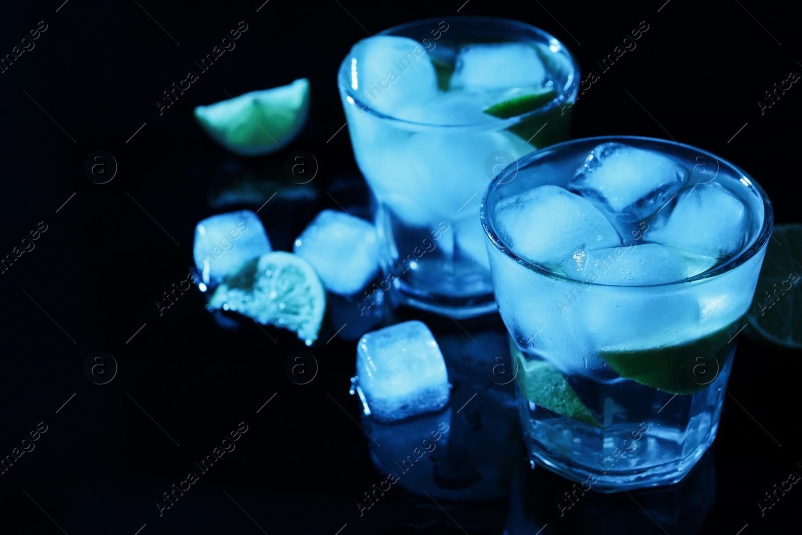 Photo of Shot glasses of vodka with ice cubes and lime slices on dark background, space for text