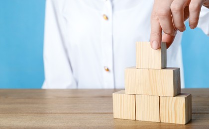 Photo of Woman building pyramid of cubes on wooden table against light blue background, closeup. Space for text