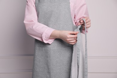 Photo of Woman putting on grey apron against white wall, closeup