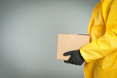 Photo of Man in chemical protective suit holding cardboard box on light grey background, closeup view with space for text. Prevention of virus spread