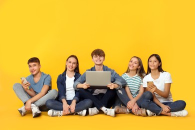 Photo of Group of happy teenagers with laptop and smartphones showing thumbs up on orange background