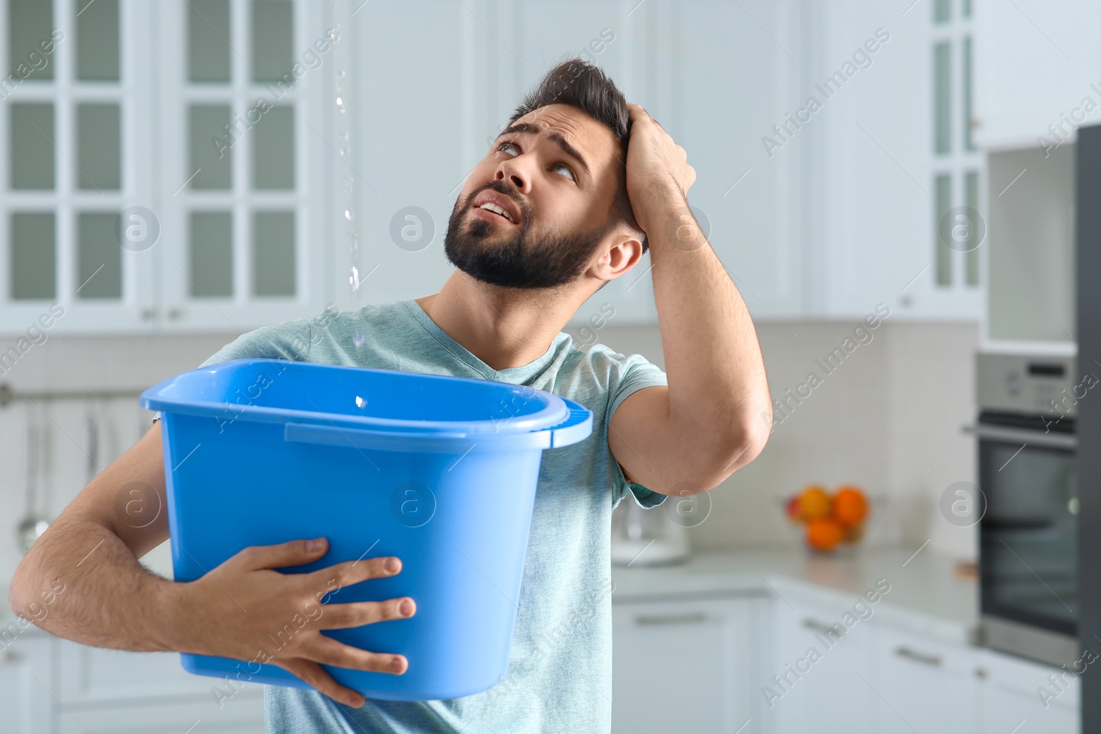 Photo of Young man collecting leaking water from ceiling in kitchen. Time to call roof repair service