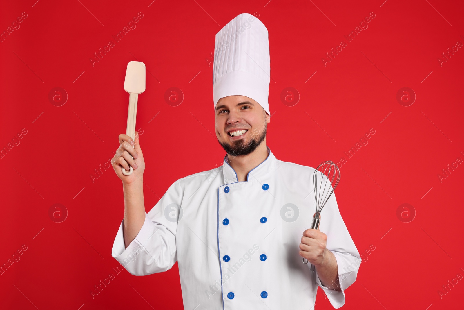 Photo of Happy professional confectioner in uniform holding whisk and spatula on red background