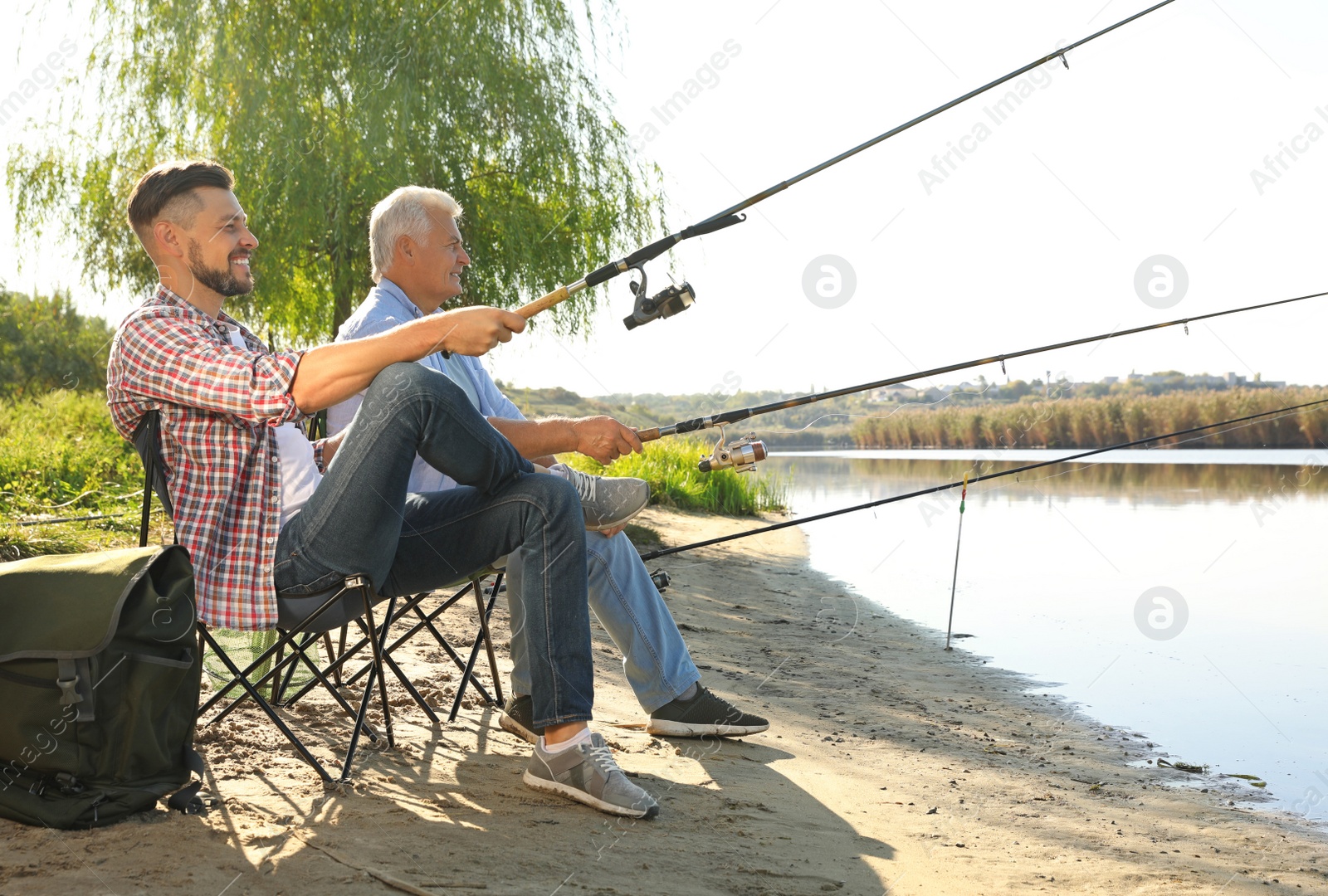 Photo of Father and adult son fishing together from riverside on sunny day