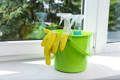 Photo of Green bucket with gloves and spray bottles of detergents on window sill indoors, space for text. Cleaning supplies
