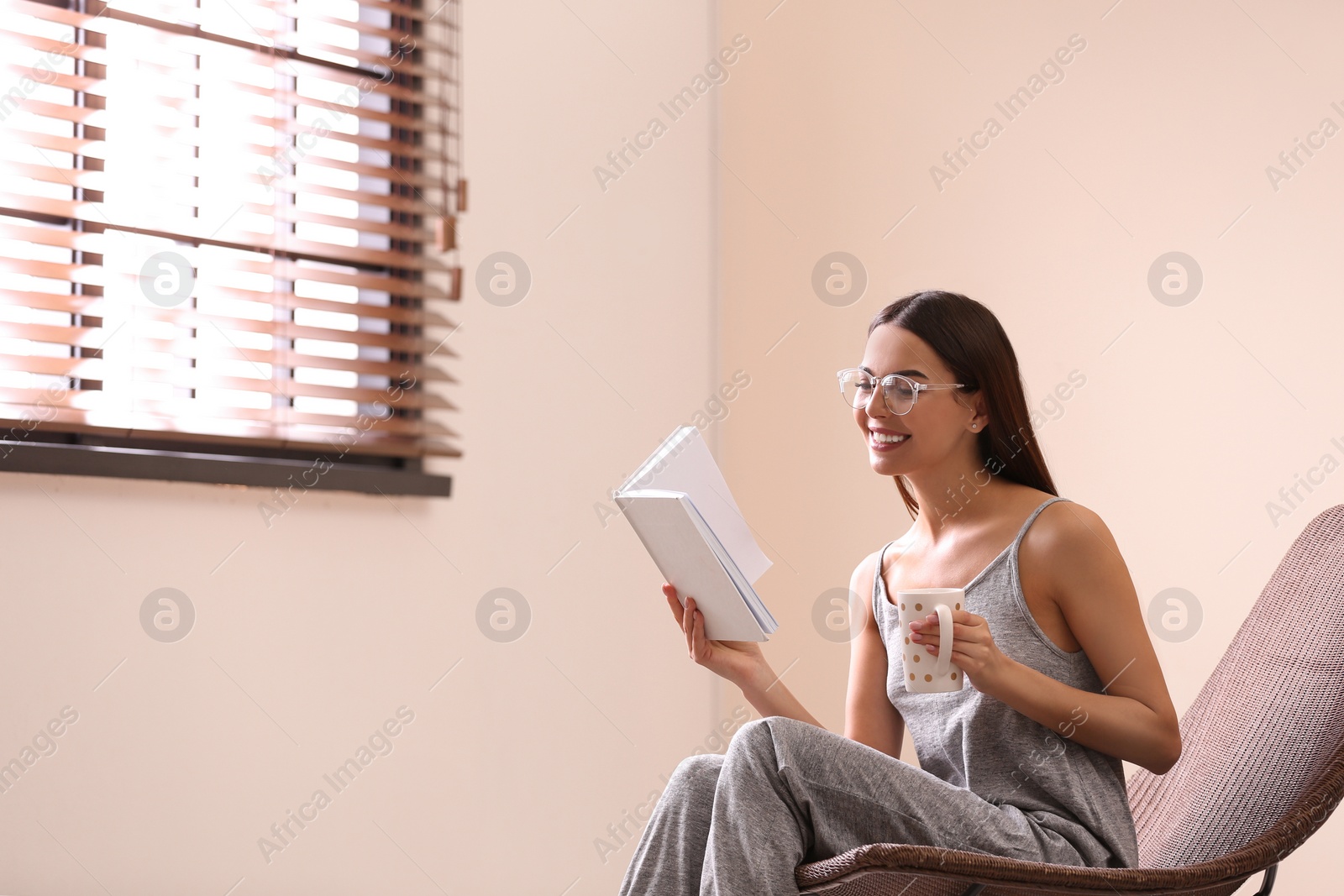 Photo of Young woman reading book near window with blinds at home. Space for text