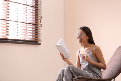 Photo of Young woman reading book near window with blinds at home. Space for text