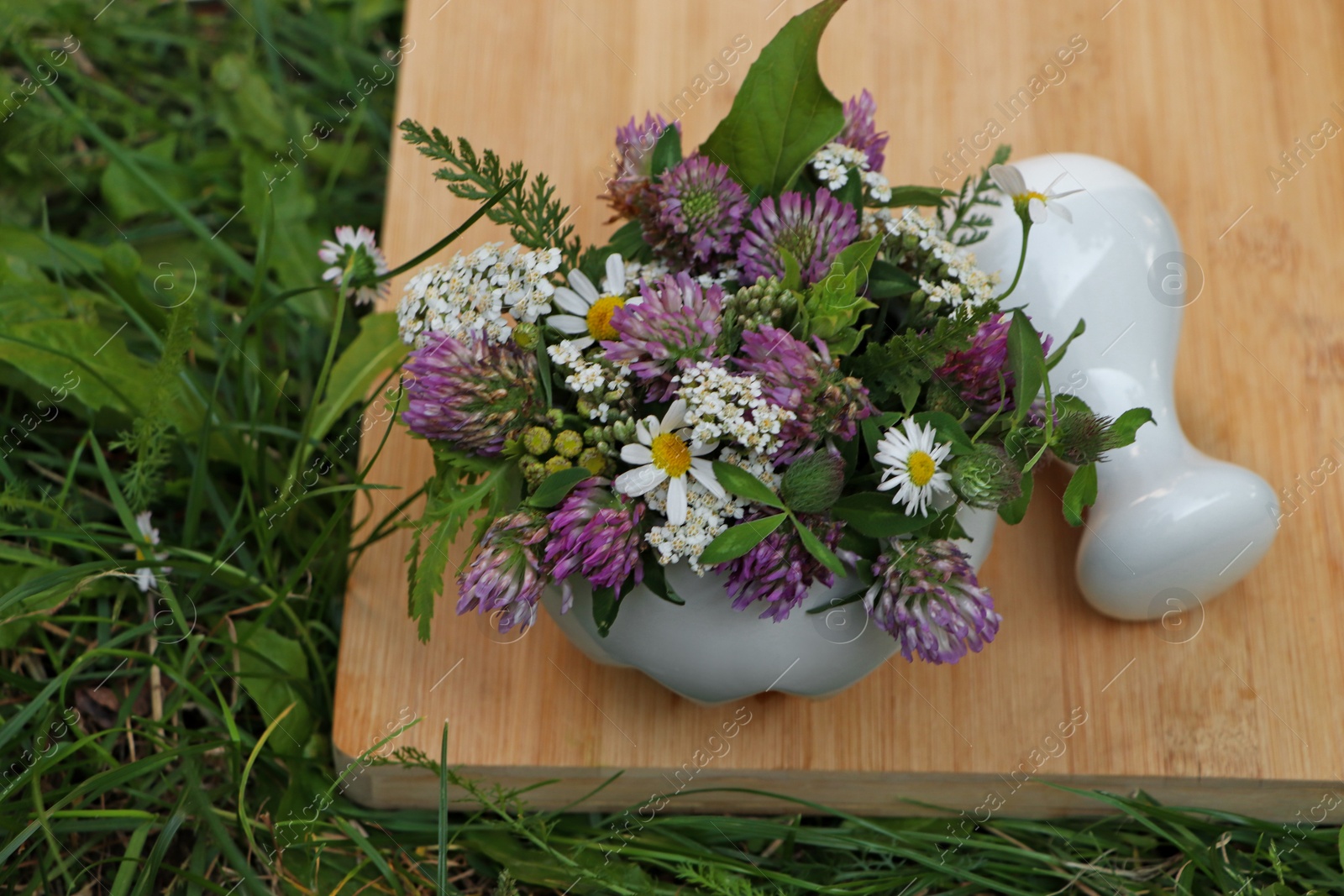 Photo of Ceramic mortar with pestle, different wildflowers and herbs on green grass, above view