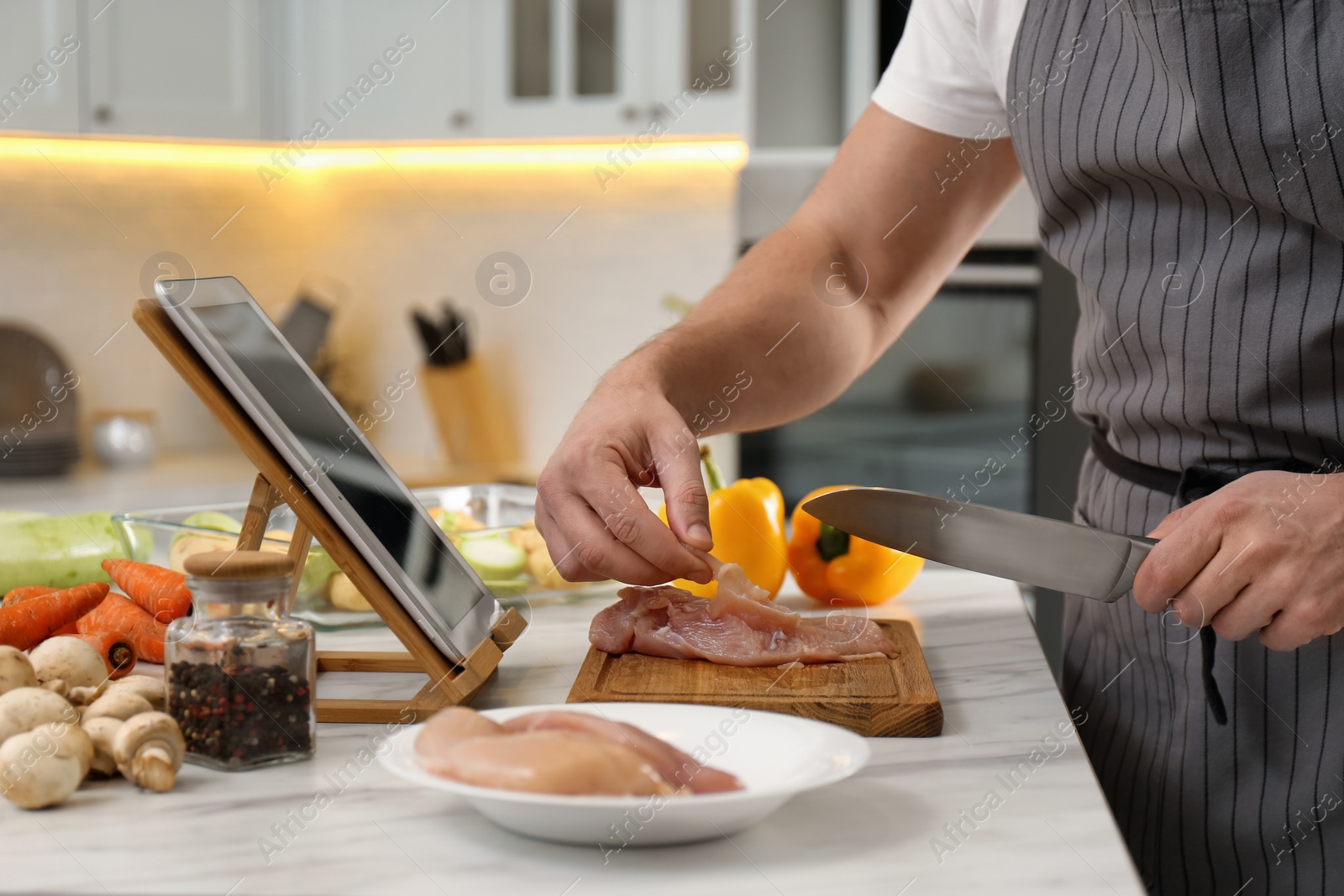 Photo of Man cutting chicken fillet while watching online cooking course via tablet in kitchen, closeup