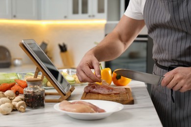 Man cutting chicken fillet while watching online cooking course via tablet in kitchen, closeup