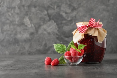 Glass jar of sweet jam with ripe raspberries and green leaves on grey table. Space for text