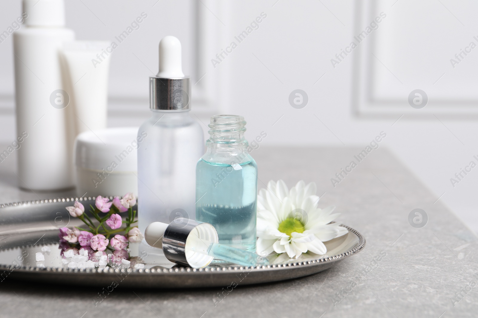 Photo of Bottles of cosmetic serum and flowers on gray table, space for text