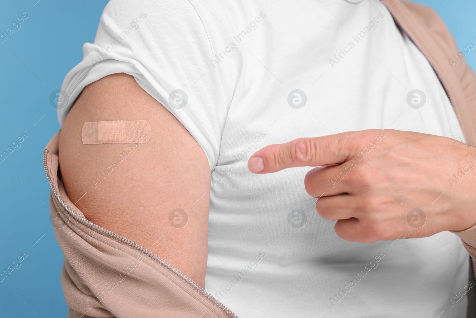 Photo of Man pointing at adhesive bandage after vaccination on light blue background, closeup