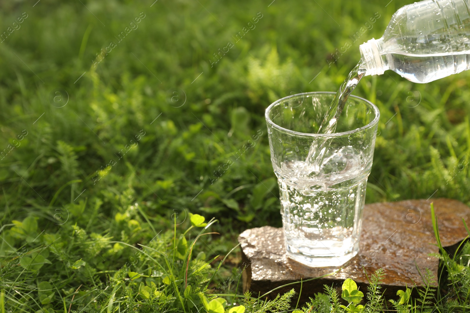 Photo of Pouring fresh water from bottle into glass on stone in green grass outdoors. Space for text