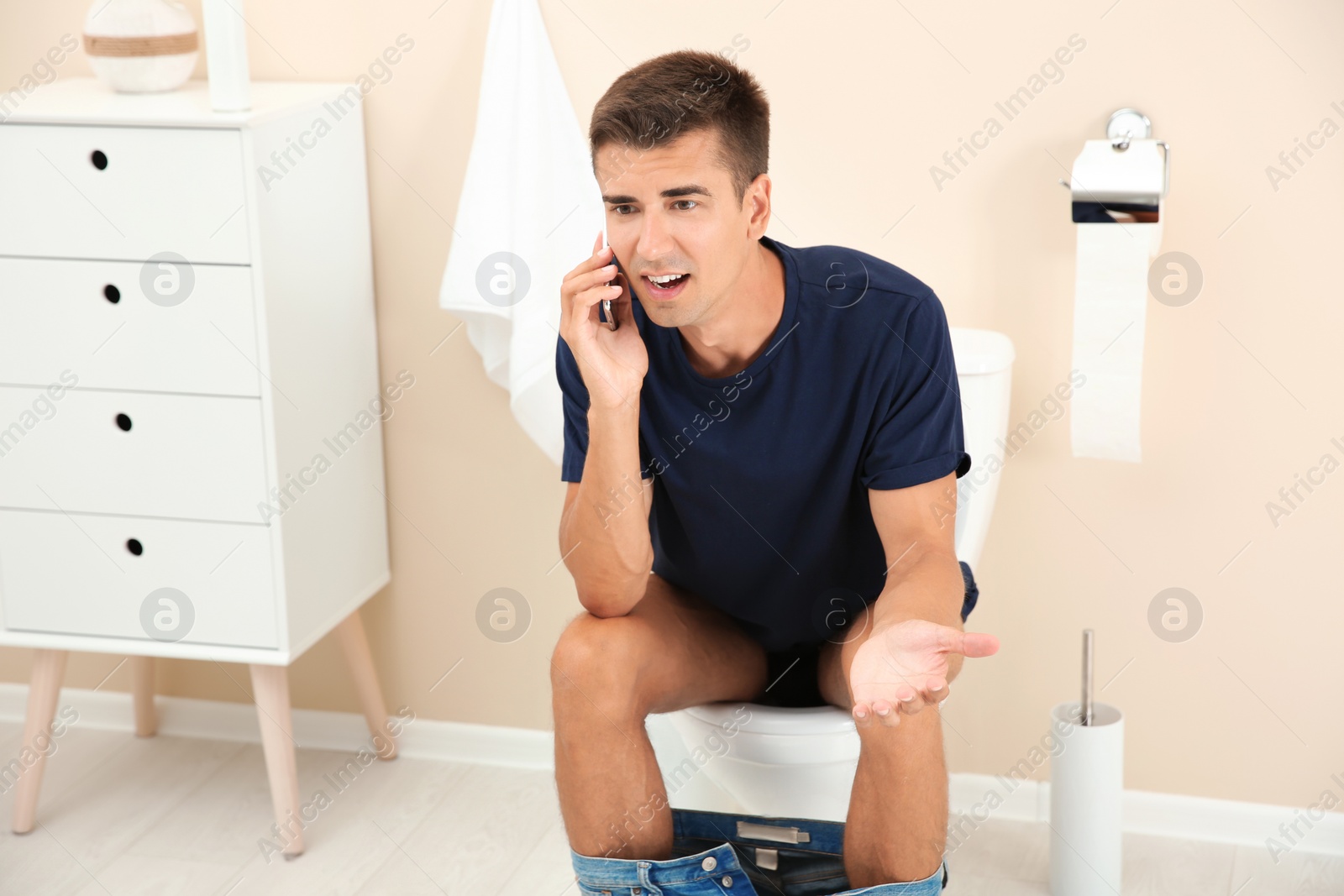 Photo of Young man with mobile phone sitting on toilet bowl at home