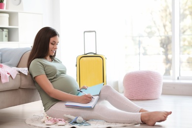 Pregnant woman making list while packing suitcase for maternity hospital in living room