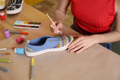 Photo of Woman painting on sneaker at wooden table indoors, closeup. Customized shoes