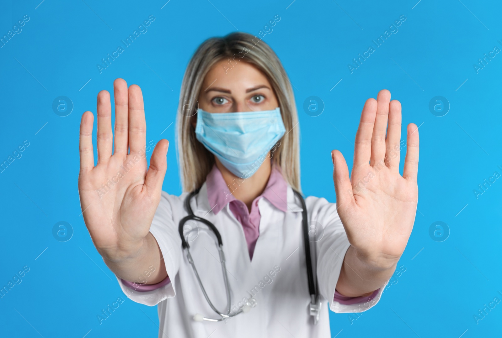 Photo of Doctor in protective mask showing stop gesture on light blue background. Prevent spreading of coronavirus