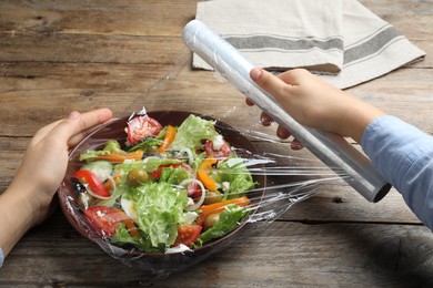 Photo of Woman putting plastic food wrap over bowl of fresh salad at wooden table, closeup