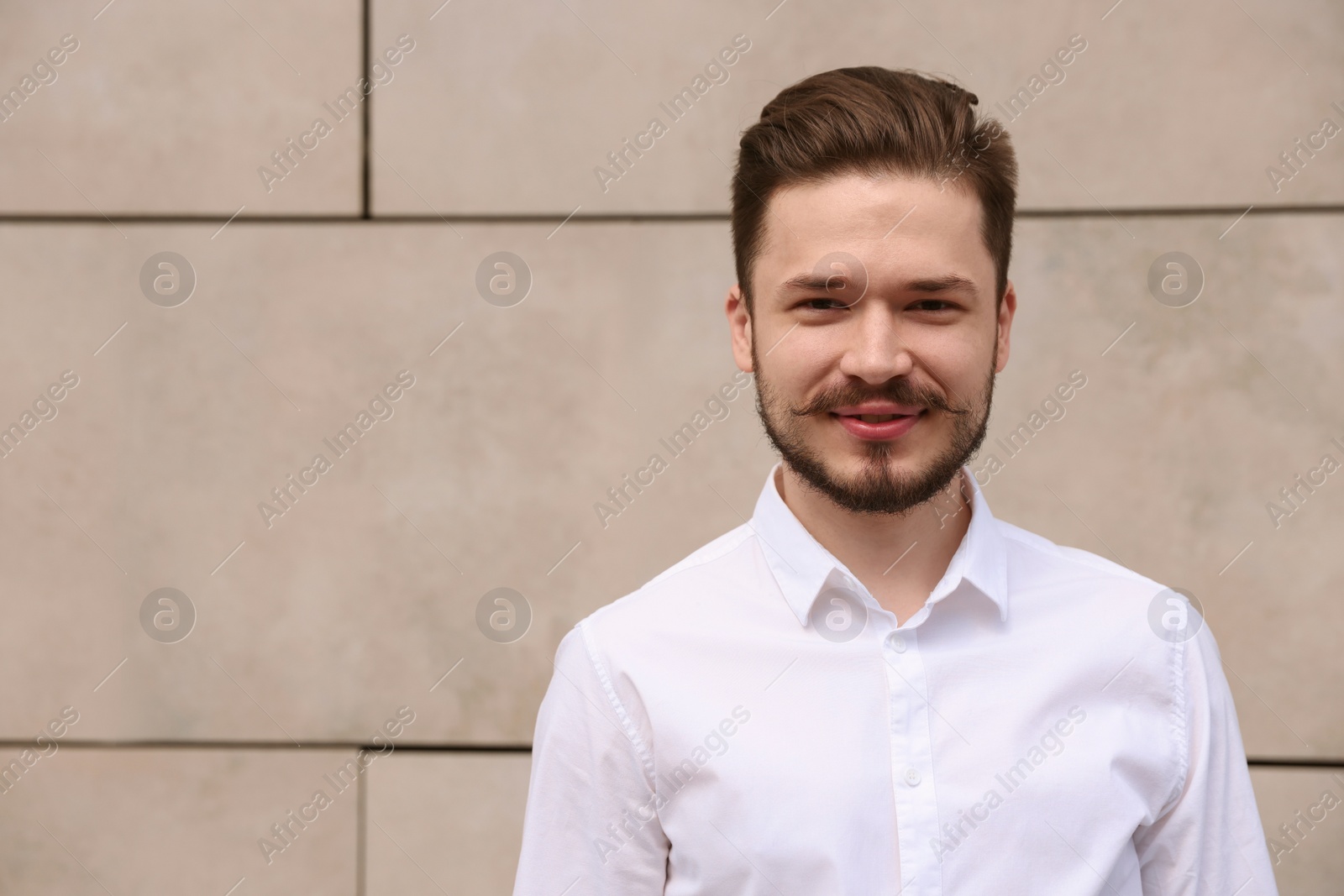 Photo of Smiling man in white shirt standing near light grey brick wall. Space for text