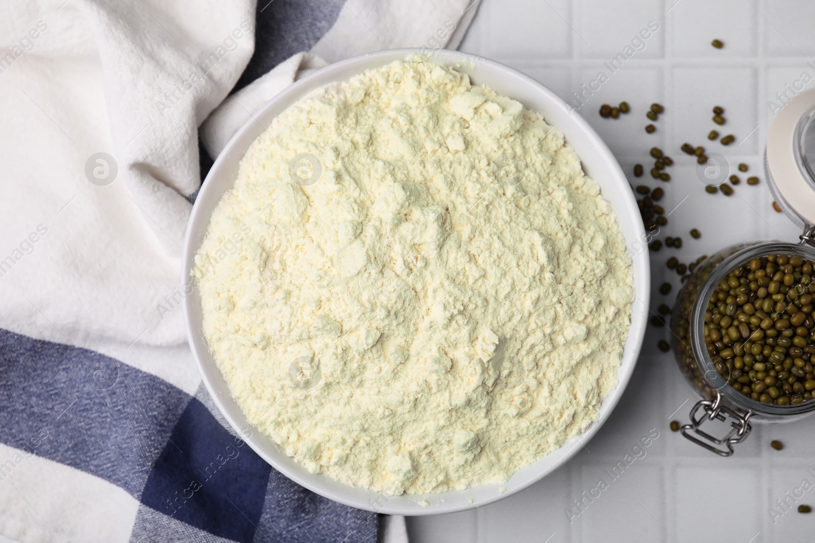 Photo of Mung bean flour in bowl and seeds on white tiled table, flat lay