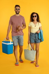 Happy young couple with cool box, bottle of beer and beach accessories on yellow background