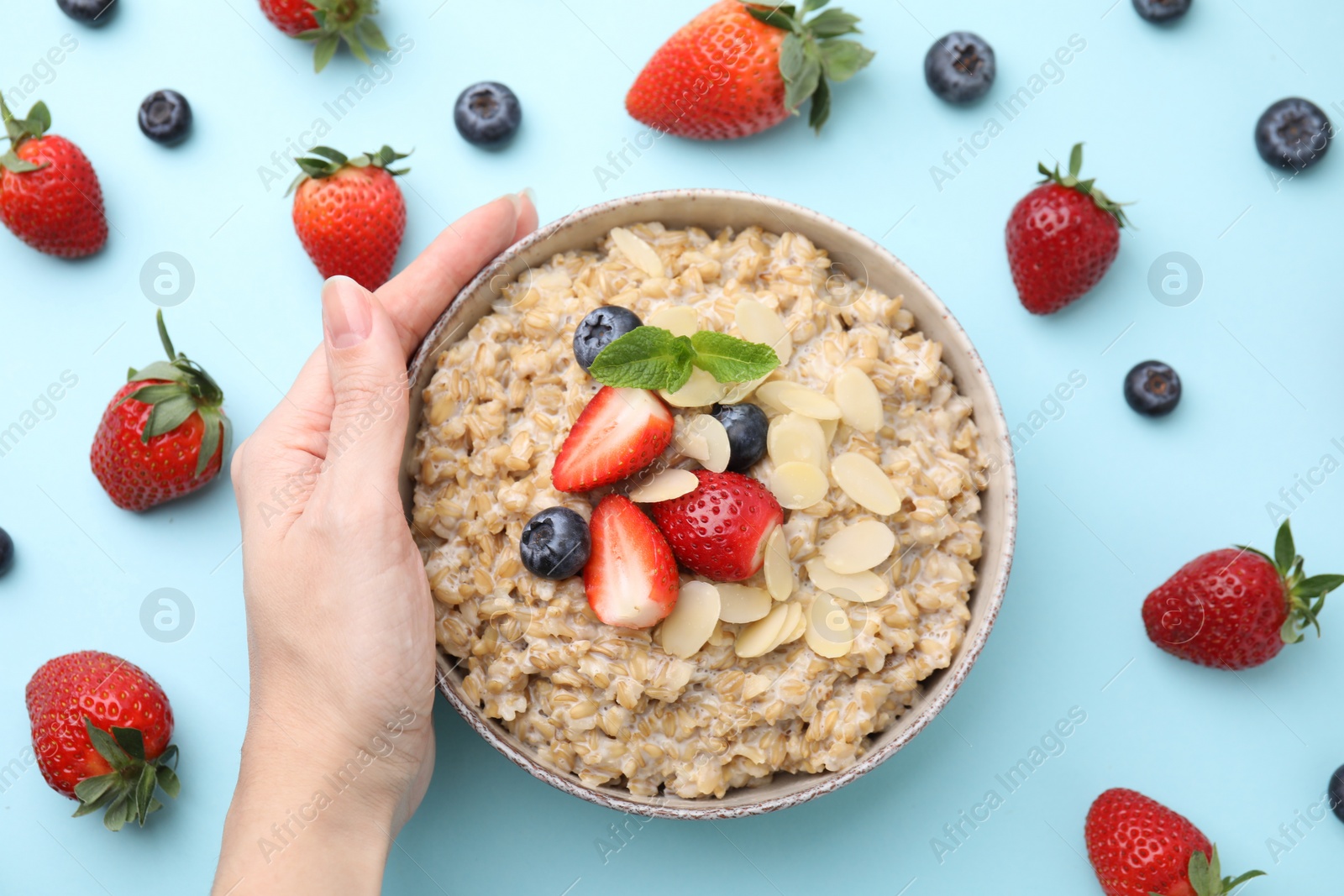 Photo of Woman holding bowl of tasty oatmeal with strawberries, blueberries and almond flakes on light blue background, top view