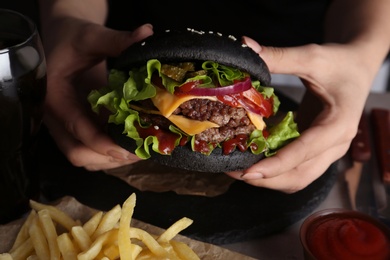 Photo of Woman holding tasty black burger at table, closeup