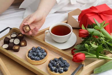 Photo of Tasty breakfast served in bed. Woman with desserts, tea, gift box and flowers at home, closeup