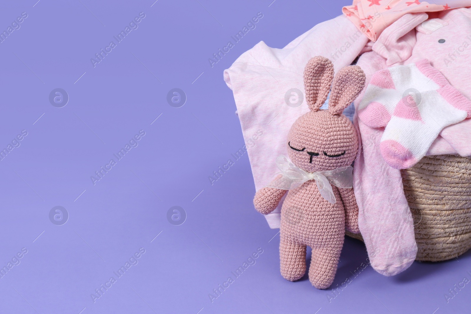 Photo of Laundry basket with baby clothes near soft toy on light purple background. Space for text