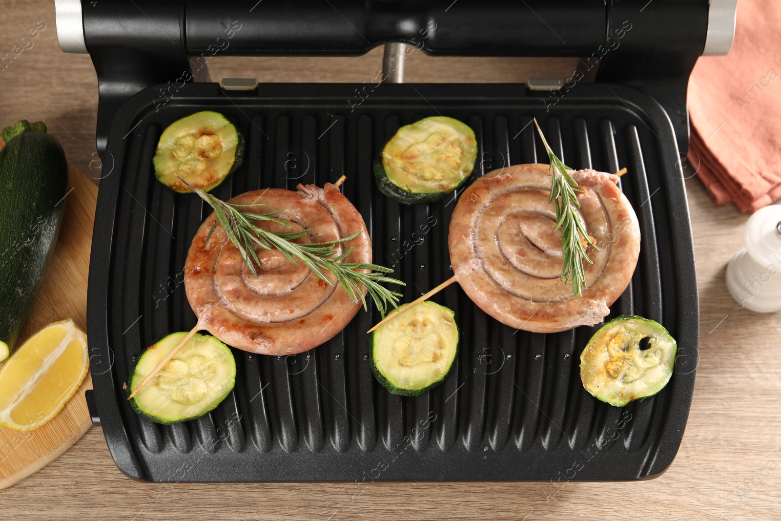 Photo of Electric grill with homemade sausages, zucchini and rosemary on wooden table, above view